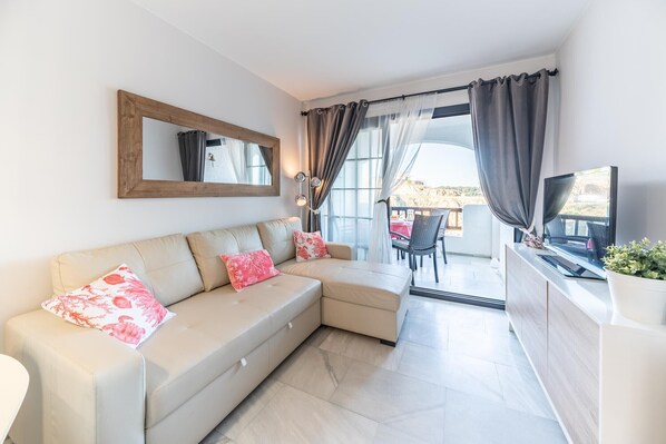 A modern living room with a beige sectional sofa, a wooden-framed mirror, a flat-screen TV, and a balcony with a view of the landscape.