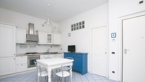 Een keuken met witte kastjes, een blauw aanrecht, een witte eettafel met stoelen, en een televisie op een blauwe kast.