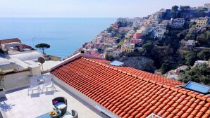 A rooftop terrace with a tiled roof, outdoor furniture, and a view of a coastal town.