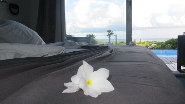 Cama com uma flor branca, vista para o mar e piscina.