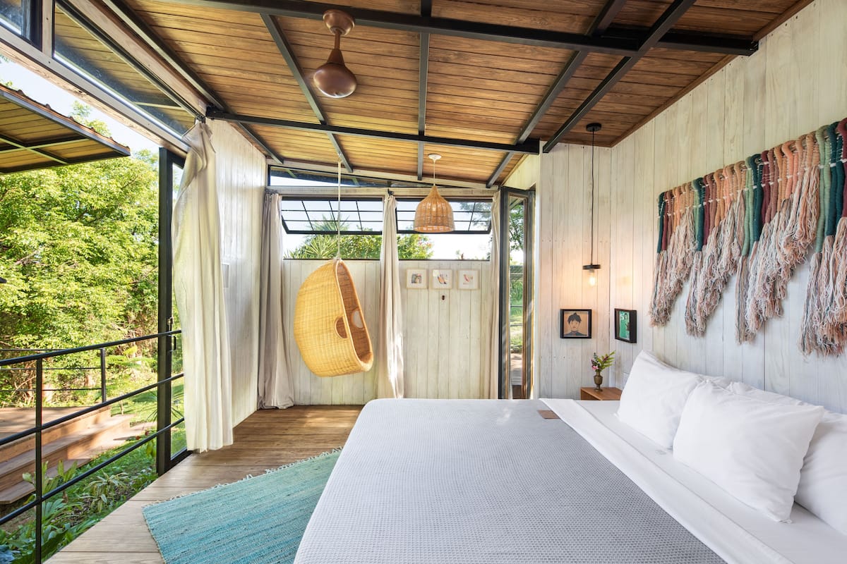 A bedroom with a large bed, wooden ceiling, hanging light fixture, and a view of greenery outside.