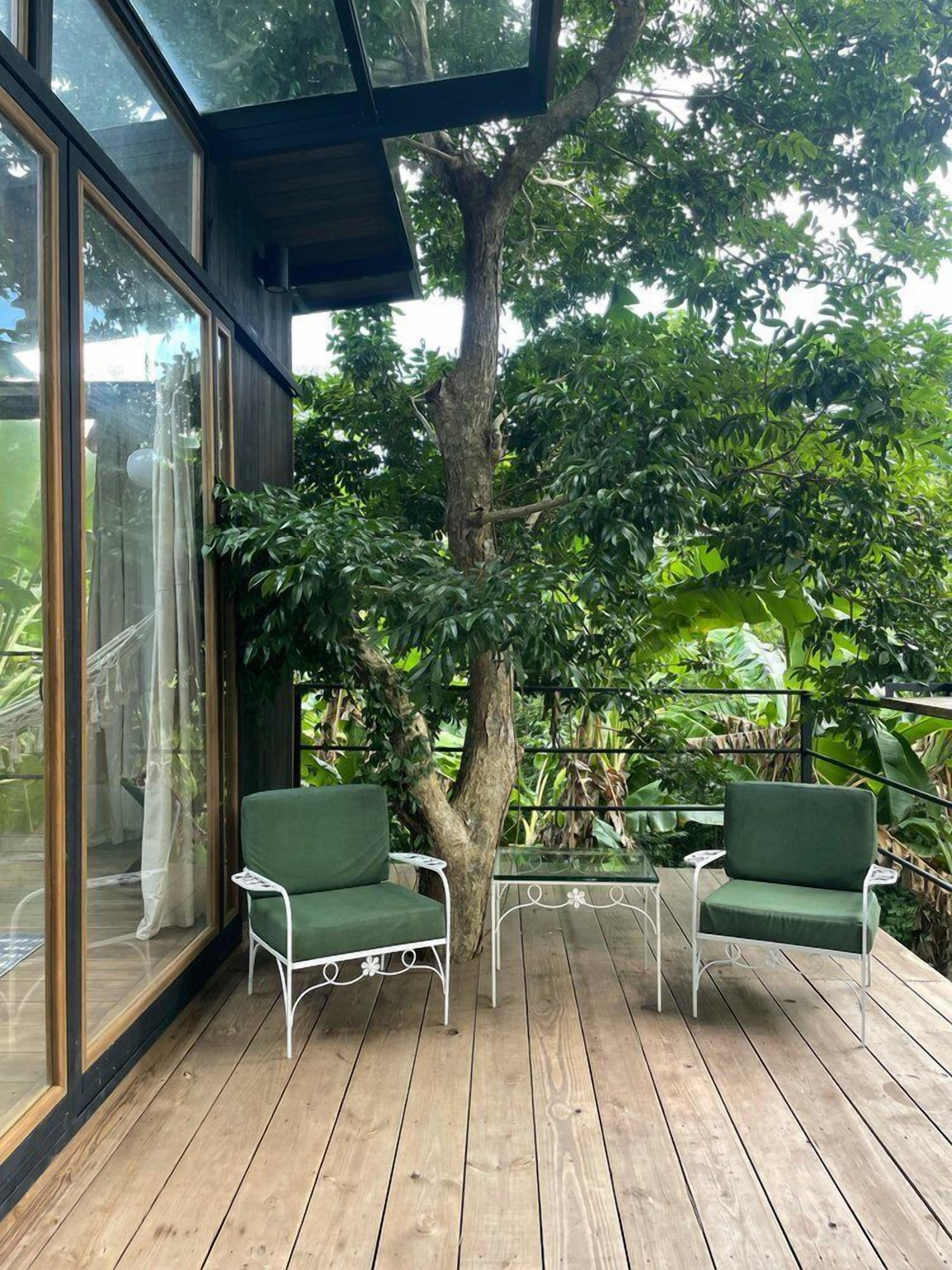 Una terraza de madera con dos sillas verdes y una mesa pequeña, rodeada de abundante follaje y un árbol grande.