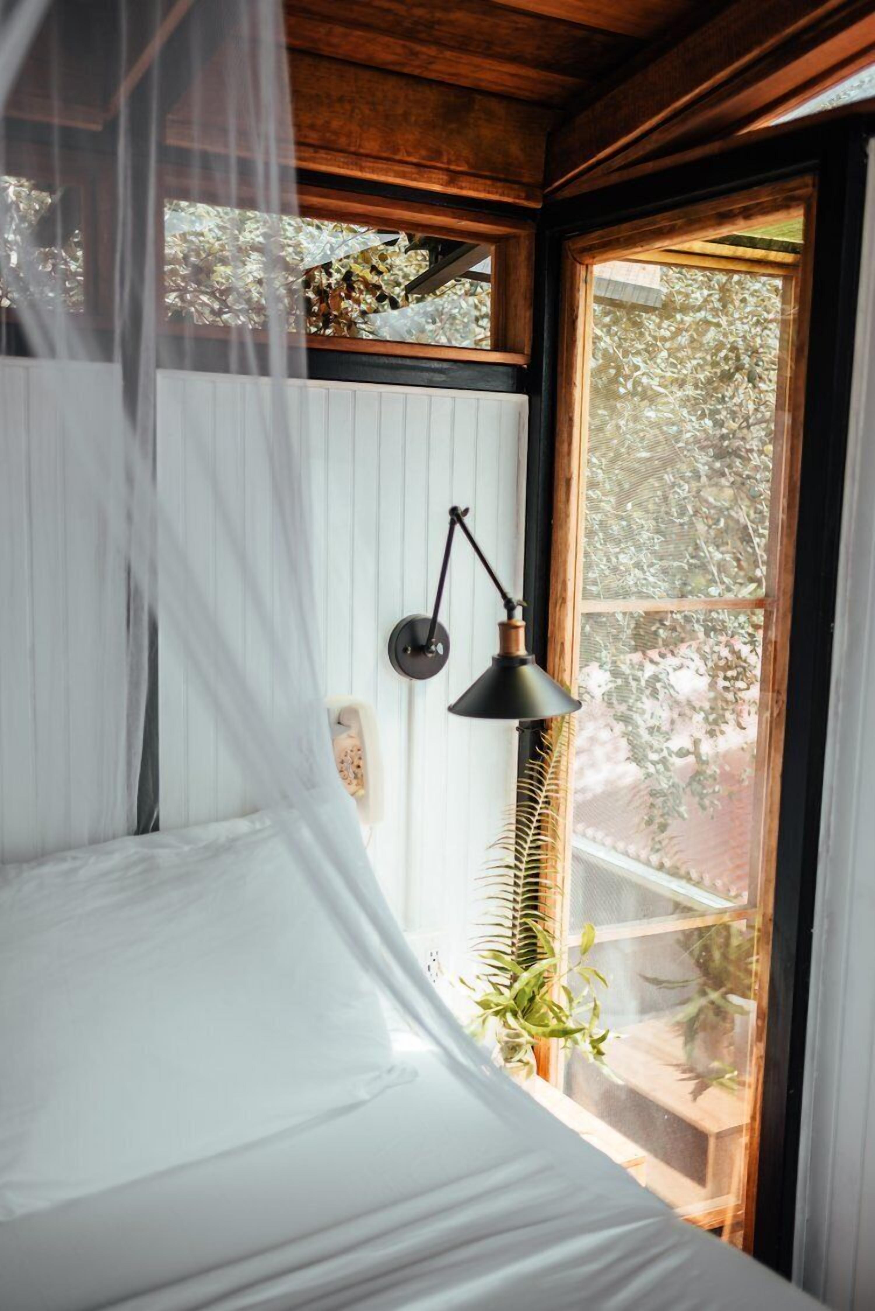 A bedroom with a canopy bed, a wall-mounted lamp, and a window with a view of greenery.