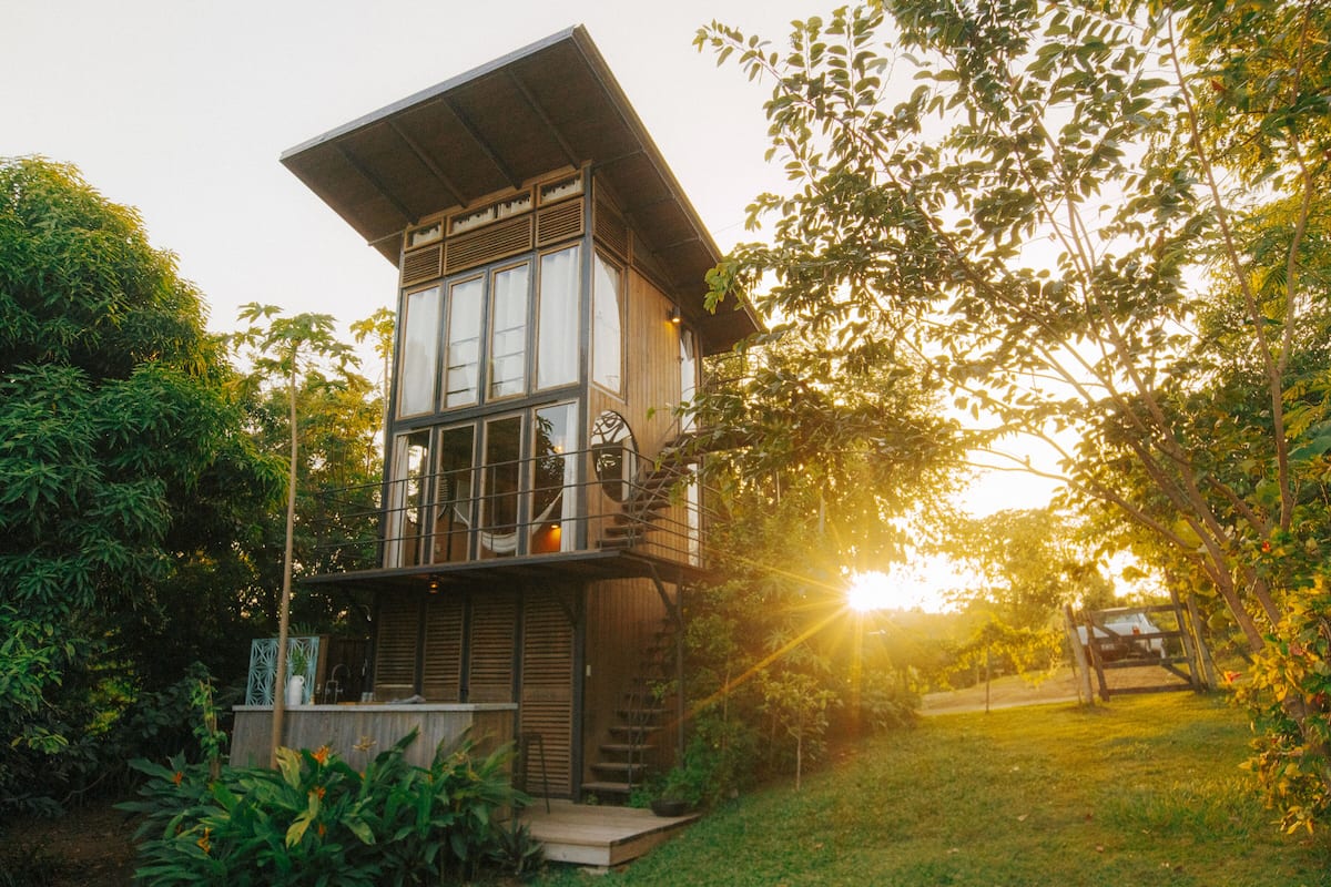 A modern two-story house with a balcony, surrounded by lush greenery and a garden.