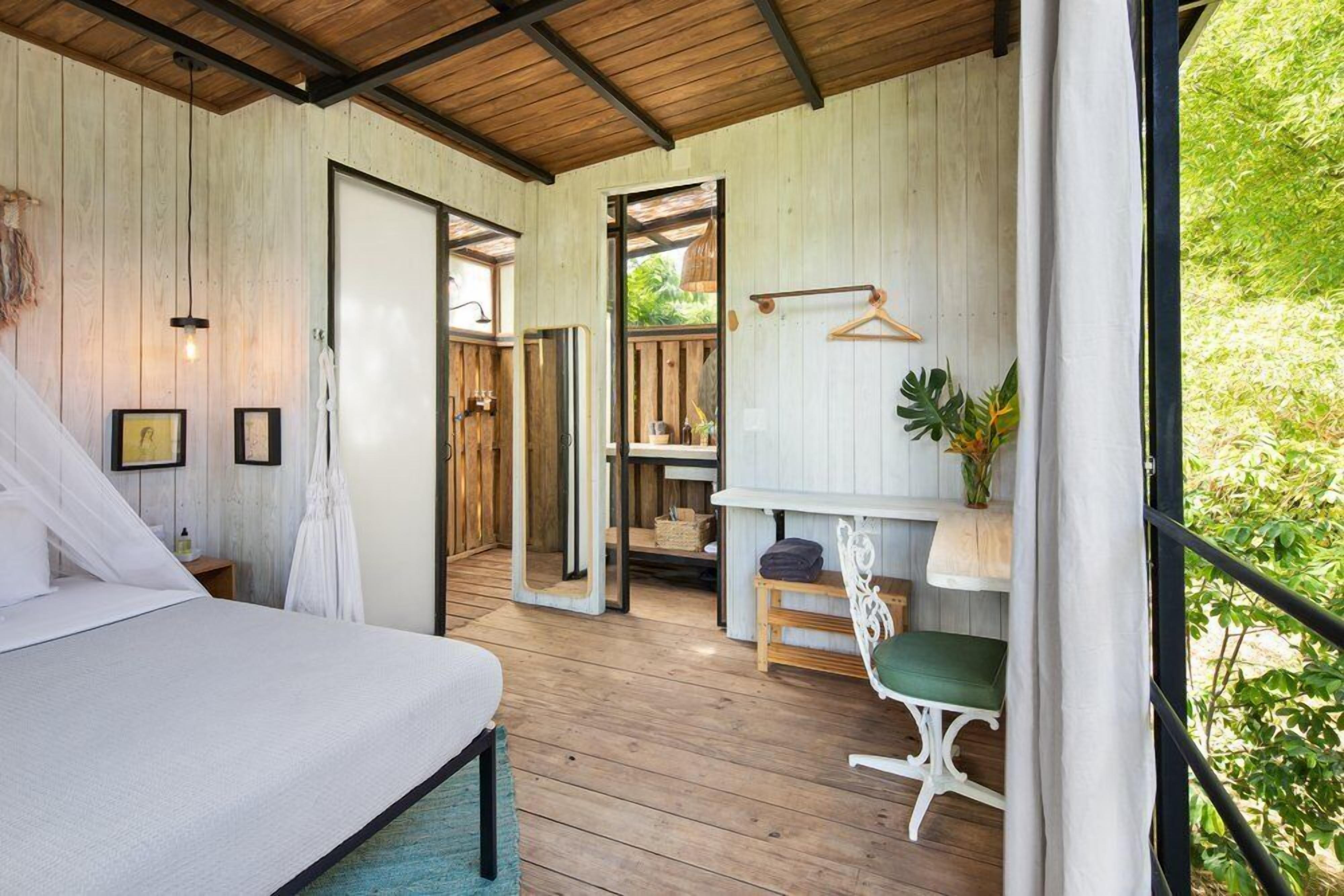 A bedroom with a bed, a desk, and a chair. There is a window with a view of greenery outside.