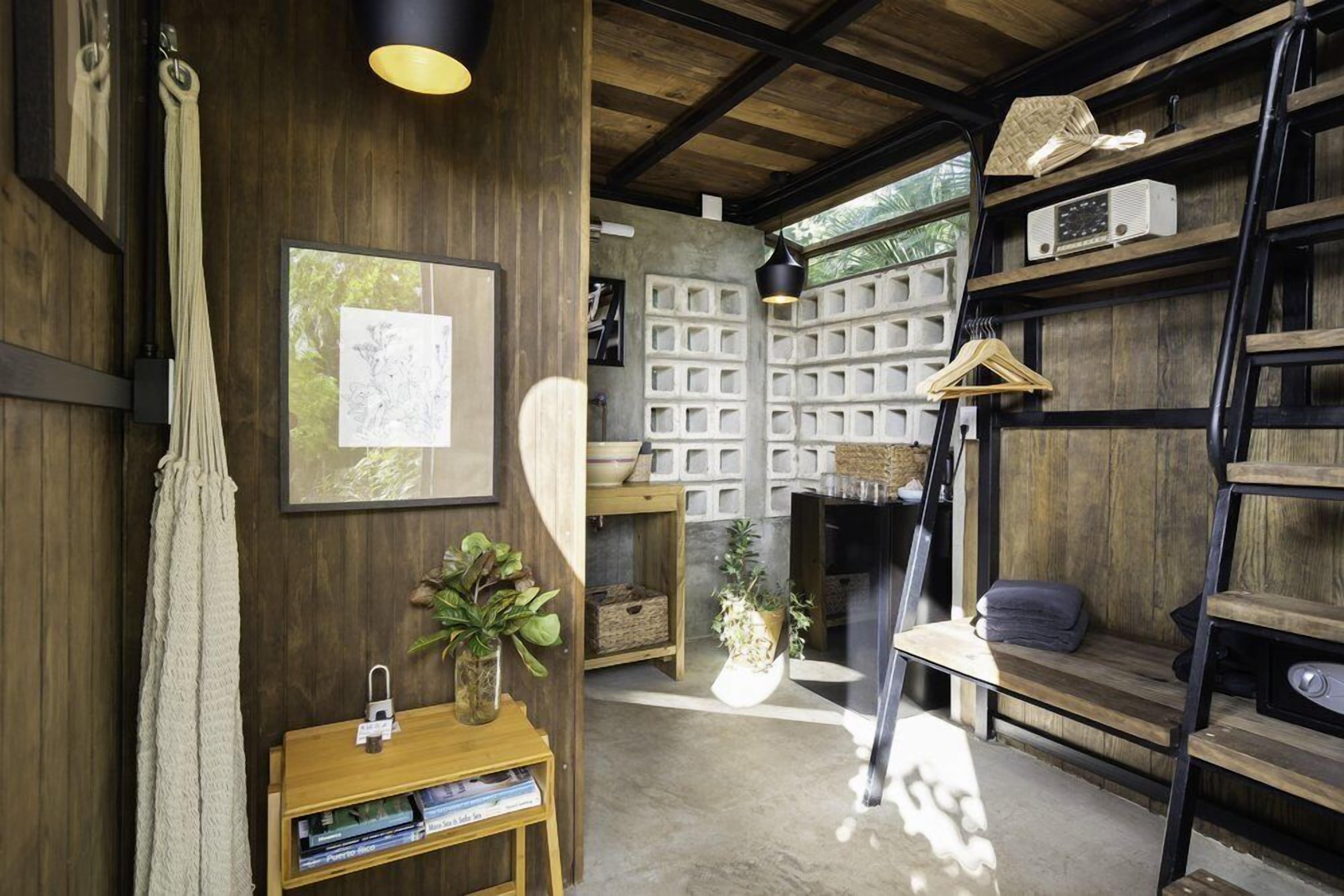 Una habitación rústica con paredes de madera, suelo de concreto y una silla colgante. Hay estantes con diversos objetos y una pequeña mesa con una planta.
