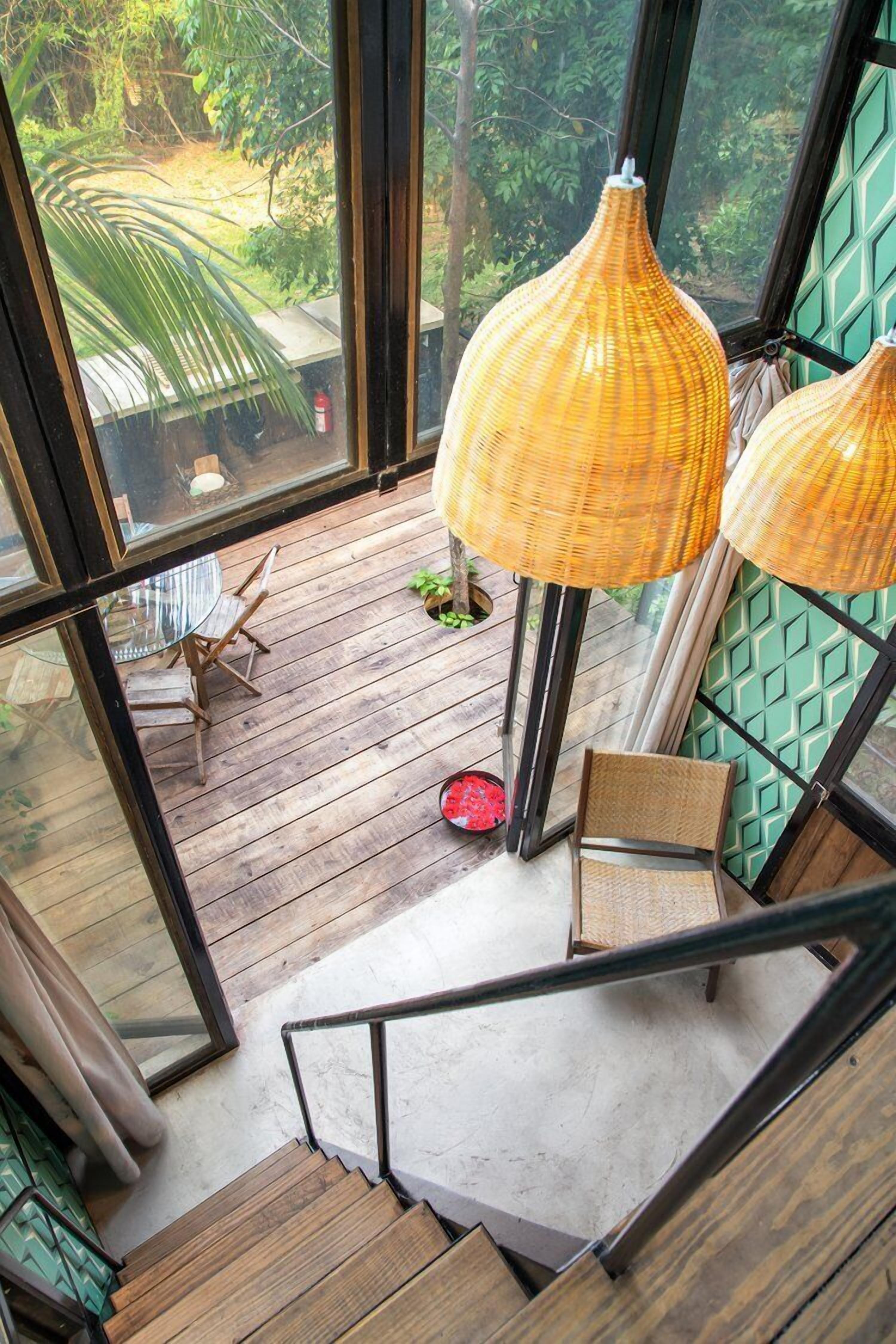 Una terraza de madera con lámparas de mimbre, una mesa de vidrio y sillas, con vistas a un jardín exuberante.