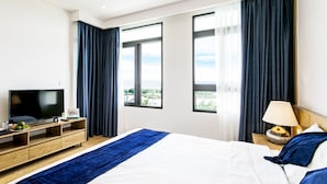 Ein Hotelzimmer mit einem großen Bett, einem Fernseher, einer Holzkommode und Blick ins Freie durch die Fenster.