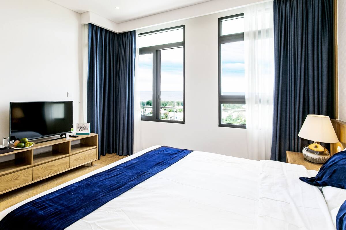 Habitación de hotel con una cama grande, televisión, cómoda de madera y vista al exterior a través de las ventanas.