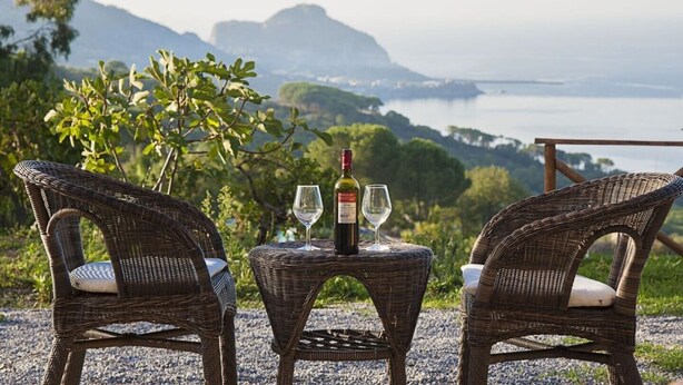 Two wicker chairs facing a table with a bottle and two wine glasses, overlooking a scenic view of a body of water and greenery. - Apartment in villa with swimming pool in Cefalù, view of the bay, App. Aranancione (Cefalu)