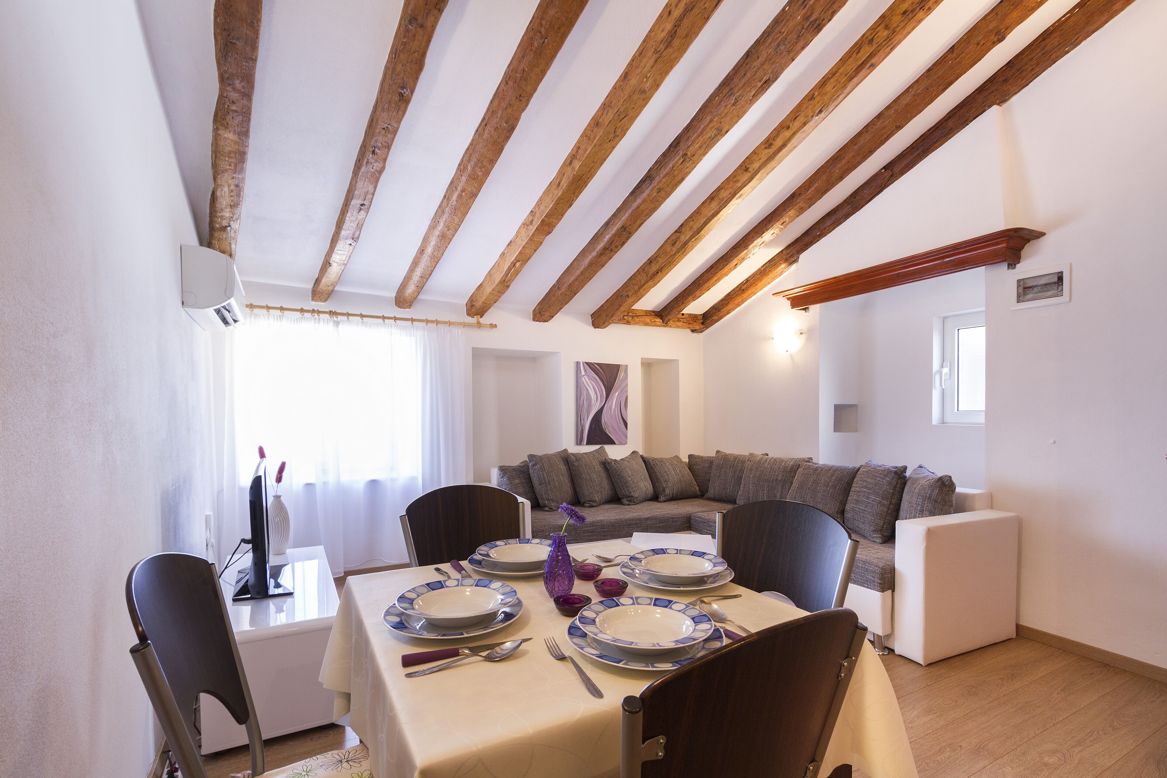 A dining area with a table set for four, wooden beams on the ceiling, a sofa, and a window with curtains.