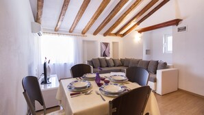 A dining area with a table set for four, wooden beams on the ceiling, a sofa, and a window with curtains.