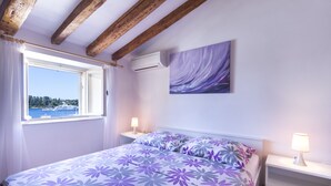 A bedroom with a bed, two bedside tables, a window with curtains, an air conditioning unit, and a painting on the wall.