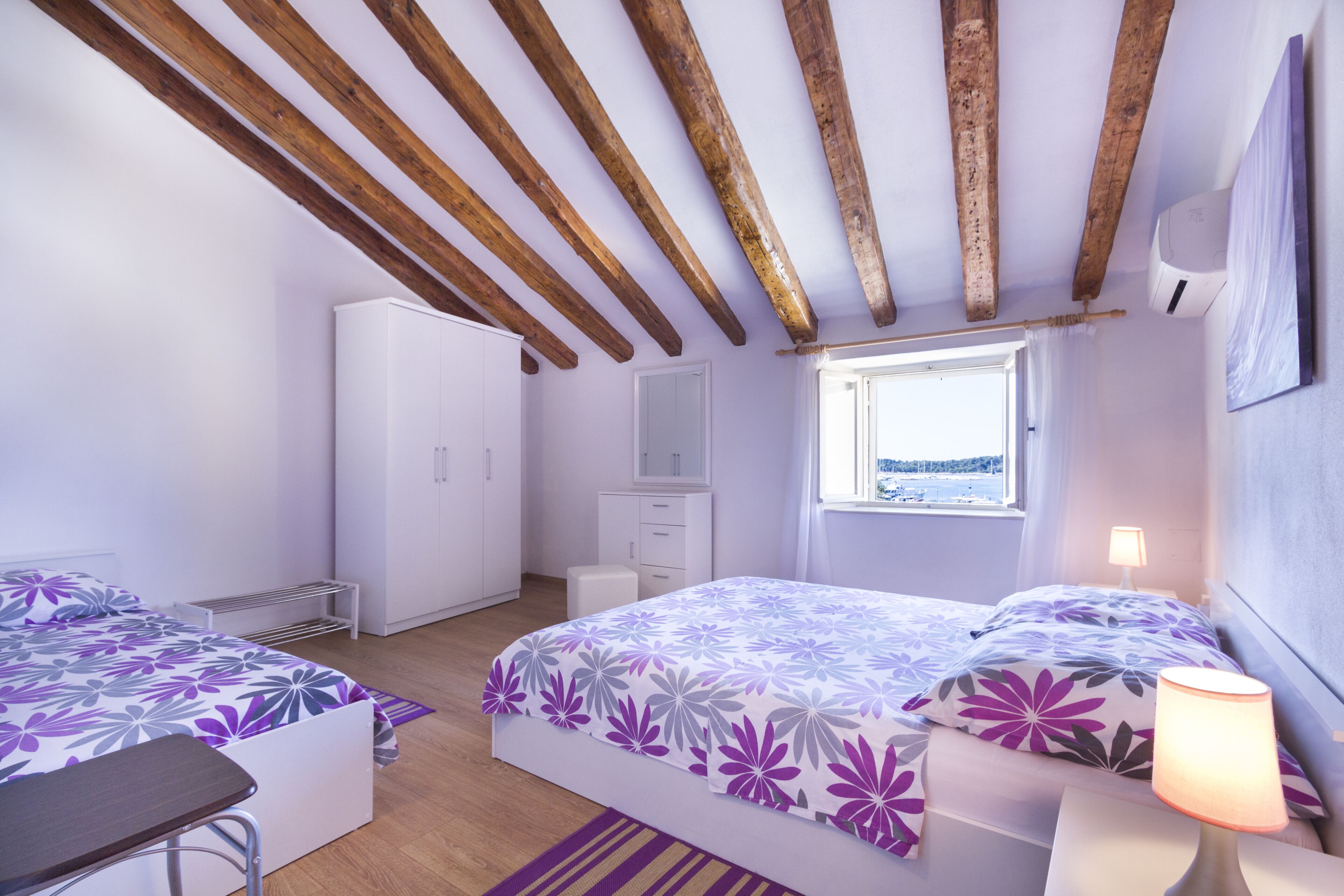 Ein Schlafzimmer mit zwei Betten, Holzbalken an der Decke, einem Fenster mit Ausblick und einem weißen Kleiderschrank.