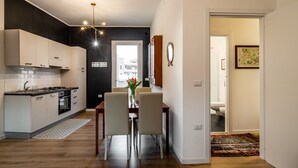 A modern kitchen with light wood cabinets, a black backsplash, and a stainless steel range hood. There is a dining area with a wooden table and white chairs, and a small rug on the floor. - La Cagliaritana - Attic in the city center  (Cagliari)