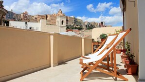A balcony with a wooden chair and a table, potted plants, and a view of buildings in the background. - La Cagliaritana - Attic in the city center  (Cagliari)