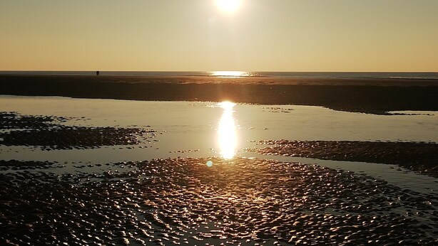 Een strandscène met een lage zon aan de horizon, die reflecteert op het natte zand.
