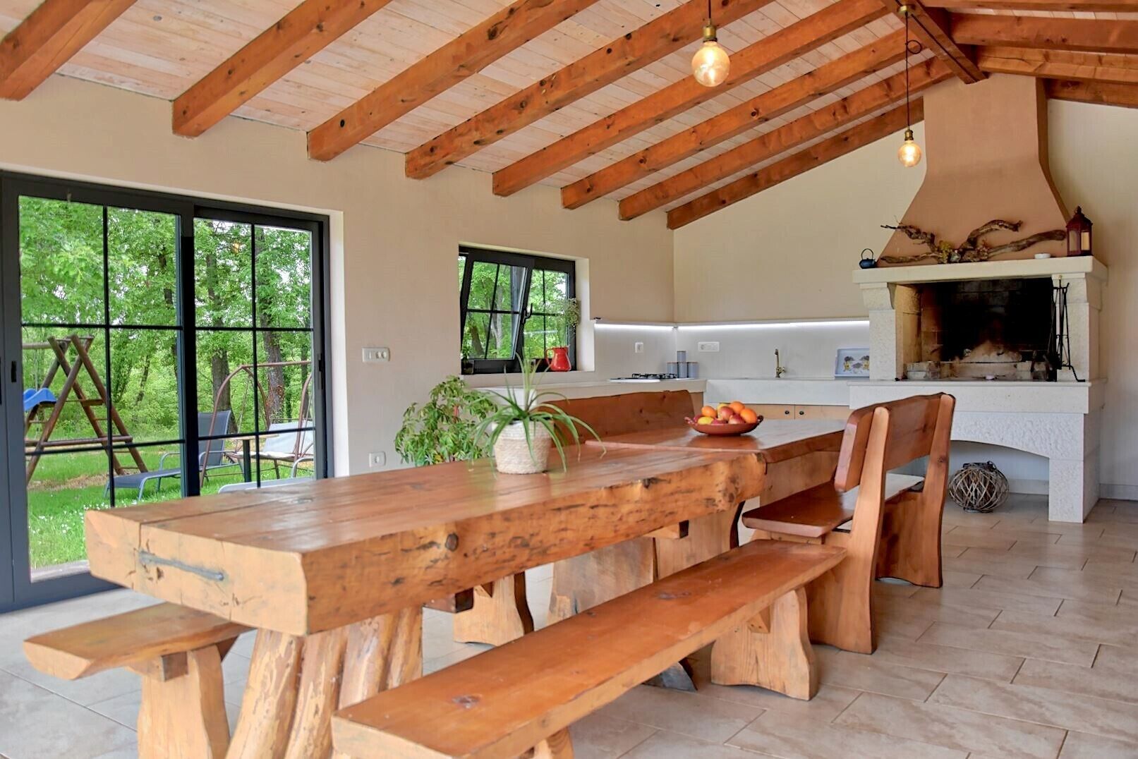 A rustic kitchen with a large wooden table and bench, a fireplace, and a view of the outdoors.