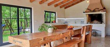 A rustic kitchen with a large wooden table and bench, a fireplace, and a view of the outdoors.