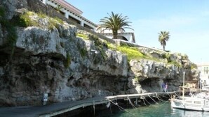 A rocky cliff with a wooden dock extending into clear water, a small boat docked, and a building with a tiled roof in the background. - Villa For Rent In Menorca With Private Pool, Enclosed Garden & Air Conditioni (Villacarlos)