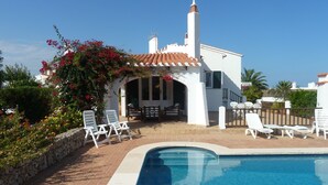 A poolside area with white lounge chairs, a stone wall, and a white building with a tiled roof. - Villa For Rent In Menorca With Private Pool, Enclosed Garden & Air Conditioni (Villacarlos)