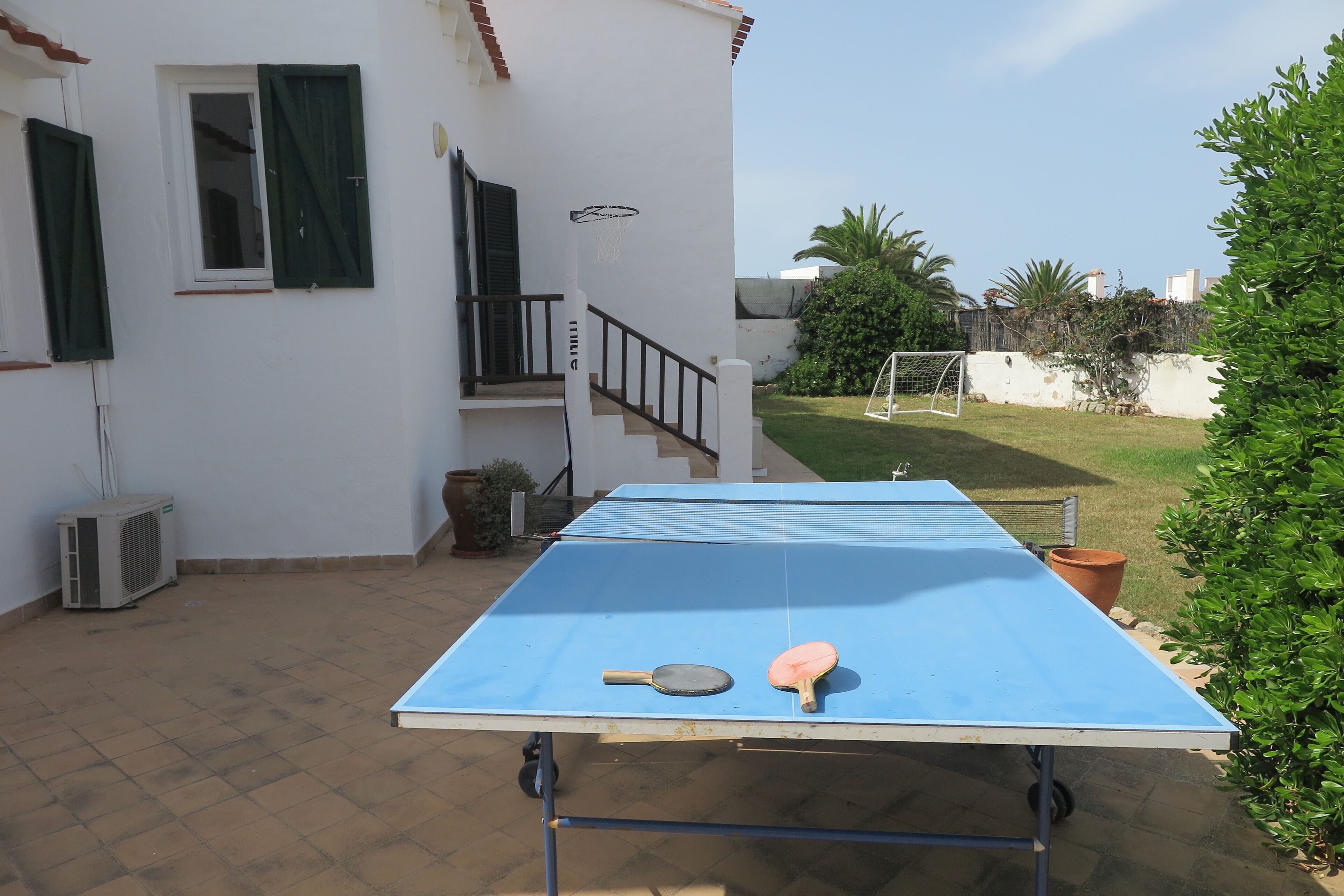 An outdoor ping pong table with two paddles, a staircase, and a white building with green shutters.