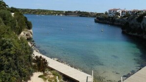 A coastal view with a clear blue sea, rocky cliffs, and a wooden walkway leading to a sandy beach. - Villa For Rent In Menorca With Private Pool, Enclosed Garden & Air Conditioni (Villacarlos)