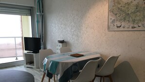 A dining area with a table covered by a striped tablecloth, surrounded by four chairs. A television is on a stand to the left, and a map is on the wall. - Fort-Mahon-Plage: Studio with balcony, fully equipped, private parking (Fort-Mahon-Plage)