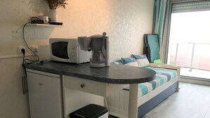 A compact kitchenette with a microwave, coffee maker, and a small fridge. A bed with striped bedding is visible in the background. - Fort-Mahon-Plage: Studio with balcony, fully equipped, private parking (Fort-Mahon-Plage)