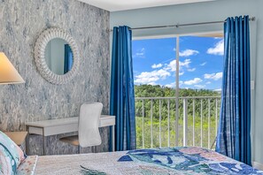 Ein Schlafzimmer mit einem großen Bett, einem Nachttisch, einem Spiegel und Blick auf einen Balkon mit blauen Vorhängen.