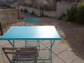 A turquoise metal table and chair set on a paved patio with a gravel path and a wooden fence in the background. - Charming Cottage in Quend-Plage: 2BR, Parking, Garden (Quend)