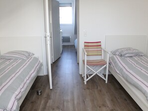 A narrow room with two single beds, a small white chair, and a door leading to another room. - Charming Cottage in Quend-Plage: 2BR, Parking, Garden (Quend)