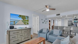A spacious living room with a wooden bench, two blue sofas, a wooden dresser, and a ceiling fan. - SEA WINDS 504 - Direct Gulf Views from Coastal Renovated Beachfront Condo! (Marco Island)