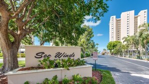 A sign for 'Sea Winds' with a large tree and flower bed in the foreground. - SEA WINDS 504 - Direct Gulf Views from Coastal Renovated Beachfront Condo! (Marco Island)
