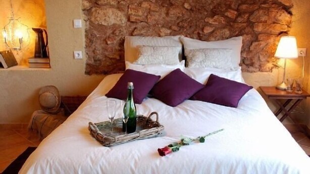 A bed with white bedding, purple pillows, a glass tray with a bottle, and a rose.