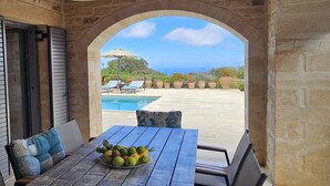 Eine überdachte Terrasse mit Esstisch, Blick auf Pool und Grünanlage, sowie eine bogenförmige Steinstruktur.
