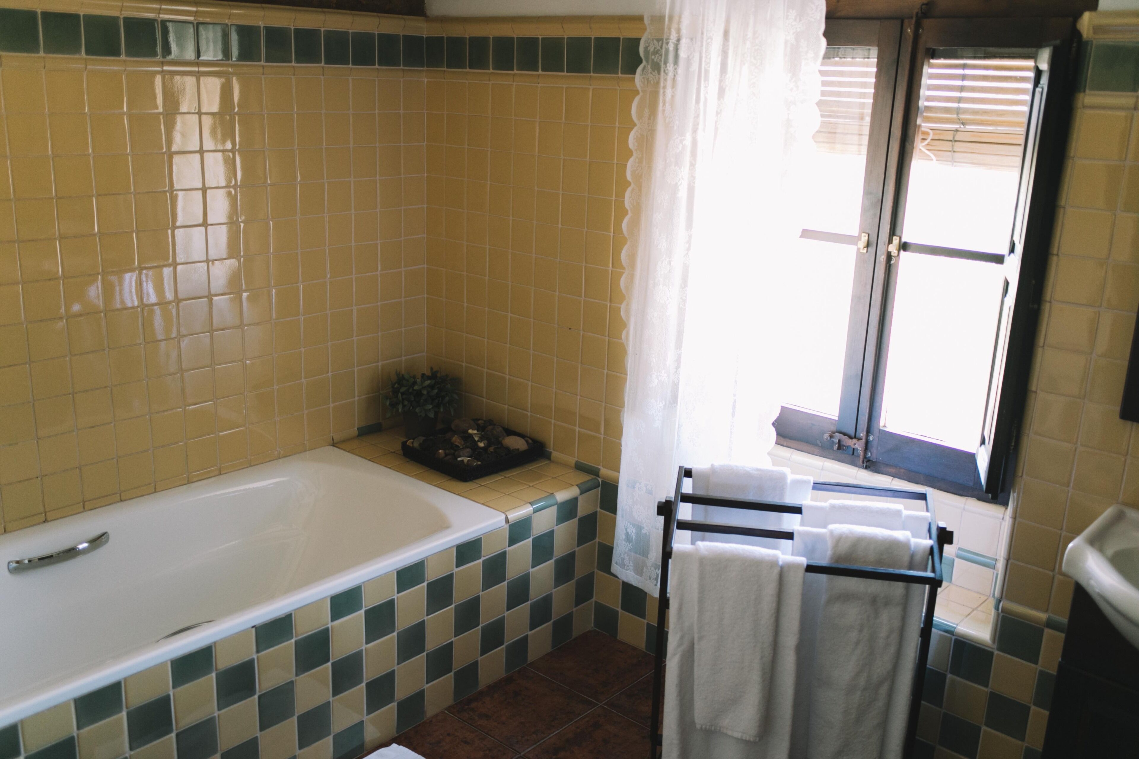 Un baño con bañera, paredes de azulejos, una ventana con persianas y un perchero con toallas.