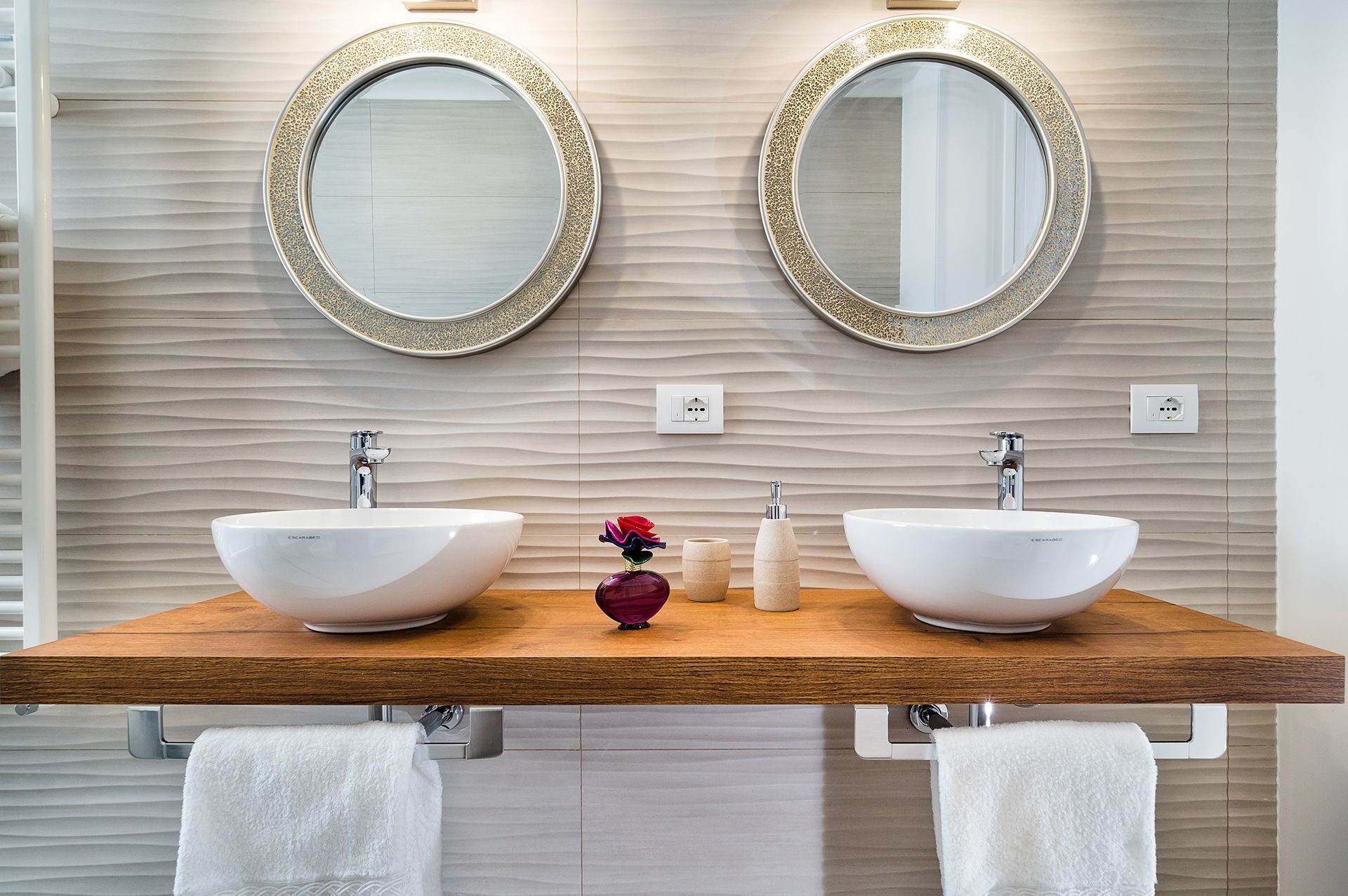 A bathroom with two white vessel sinks, a wooden vanity, and two round mirrors with gold frames.
