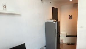 Un espace de vie compact avec un téléviseur à écran plat posé sur une console blanche, un réfrigérateur gris, un lit rouge et un plancher en bois.