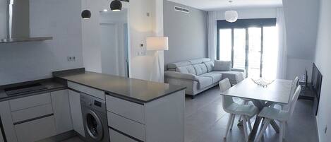 A modern kitchen with a built-in washing machine, a dining area with a table and chairs, and a living area with a sofa and a TV.