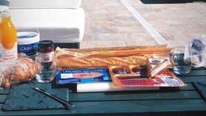 A rooftop terrace with a white sofa, lounge chairs, and a table with breakfast items like bread, butter, and a bottle of orange juice.