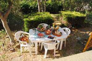 An outdoor dining table with white chairs, surrounded by greenery and trees.