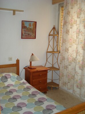 A bedroom with a bed, a nightstand, a lamp, a wooden chair, a ladder, a painting, and a window with floral curtains. - Apartment in Punta Umbria. In front of the Ria de Huelva and Muelle de las Canoas. (Punta Umbría)
