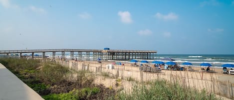 Una playa con un muelle de madera que se adentra en el océano, rodeada de sillas y sombrillas.