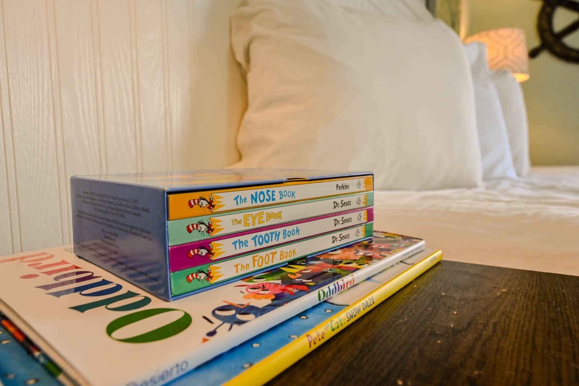 A stack of children's books on a table in a room with a bed and pillows in the background.