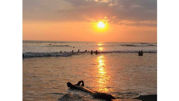 A beach scene with the sun setting over the ocean, people swimming and surfing, and a surfboard in the foreground. - Country meets beach! Las Palmitas Costa Rica (Palmar Norte)