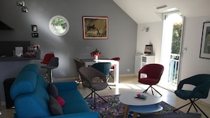 A modern living room with a blue sofa, grey armchairs, and a small round table. There is a kitchen area with a fridge and a dining table with chairs. A large window provides natural light and a view of greenery outside.