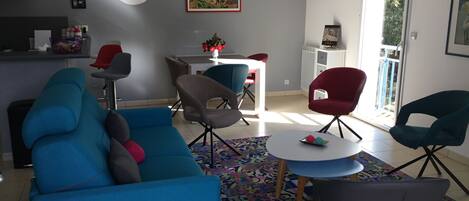 A modern living room with a blue sofa, grey armchairs, and a small round table. There is a kitchen area with a fridge and a dining table with chairs. A large window provides natural light and a view of greenery outside.