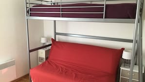 A bunk bed with a red mattress and pillows, a bedside table with a lamp, and a small drawer unit.