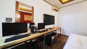 Ein Hotelzimmer mit einem Holztisch, zwei Monitoren, einer Tastatur, einer Kaffeemaschine und einem Bett.